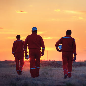 Three oil and gas workers in orange coveralls walking across a field at sunset.