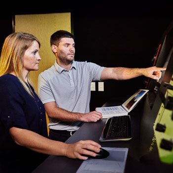 Two colleagues working together at a computer workstation, with one person pointing at a large screen.