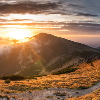 Mountain landscape at sunset with sunlight streaming through clouds and illuminating grassy slopes.