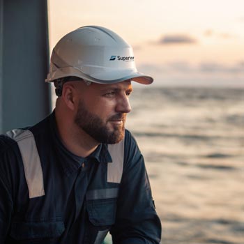 Offshore worker wearing a white hard hat and coveralls standing near railing with the ocean and sunset in the background.
