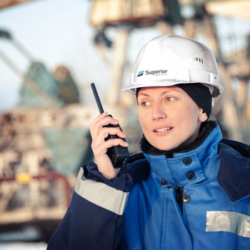 Oilfield worker in a blue jacket and white hard hat using a walkie-talkie on site.