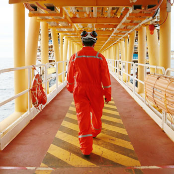 Offshore oil rig worker in orange coveralls and safety gear walking along a yellow-striped walkway on a platform.