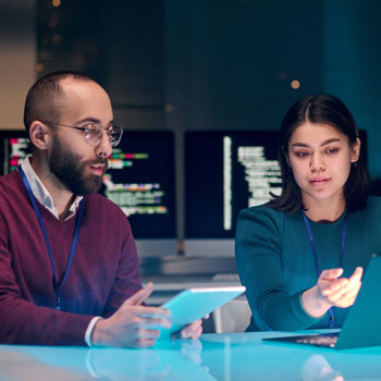 Two professionals collaborating at a desk with digital tablets and computer screens displaying code in the background.