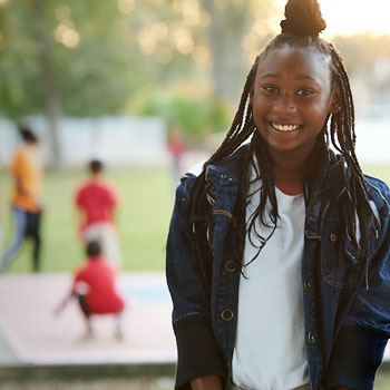 Young african american girl wearing a denim jacket and white shirt standing outdoors with children playing in the background.