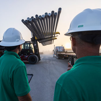 Two workers in green shirts and white hard hats observing a forklift transporting metal pipes at an industrial site during sunrise or sunset.